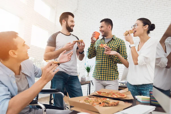 Trabalhadores de escritório e homem em uma cadeira de rodas estão comendo pizza. Eles trabalham em um escritório brilhante e moderno . — Fotografia de Stock