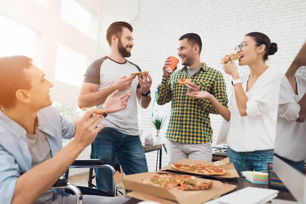 Kontorarbeidere og menn i rullestol spiser pizza. De jobber på et lyst og moderne kontor. . – stockfoto