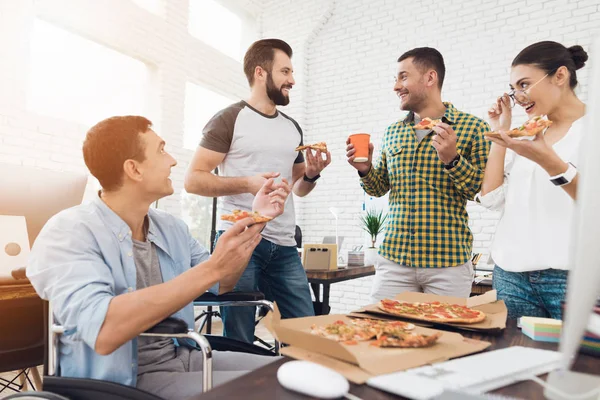 Trabalhadores de escritório e homem em uma cadeira de rodas estão comendo pizza. Eles trabalham em um escritório brilhante e moderno . — Fotografia de Stock