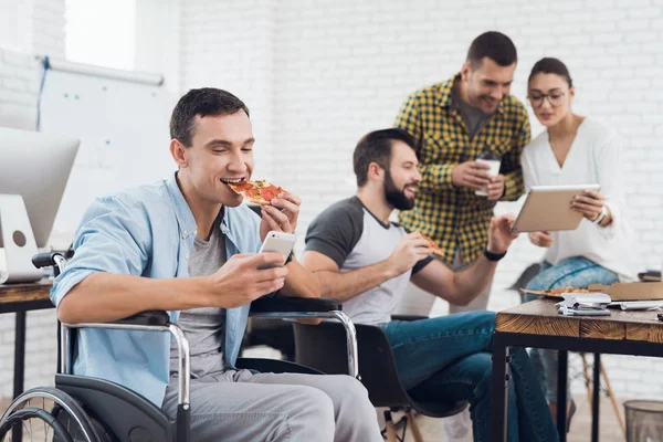 Ein Mann im Rollstuhl blickt auf den Smartphone-Bildschirm und isst ein Stück Pizza. — Stockfoto