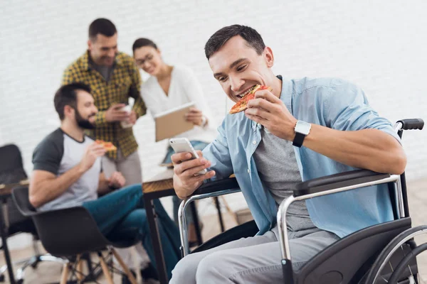 Un uomo su una sedia a rotelle guarda lo schermo dello smartphone e mangia un pezzo di pizza . — Foto Stock