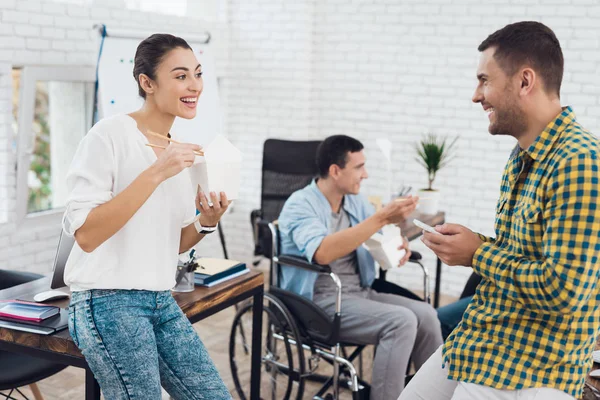 En gruppe unge og menn i rullestol snakker og spiser thaimat. De er på et lyst kontor. – stockfoto