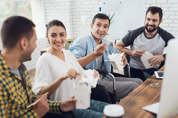 Um grupo de jovens e homens em cadeira de rodas estão conversando e comendo comida tailandesa. Eles estão em um escritório brilhante — Fotografia de Stock