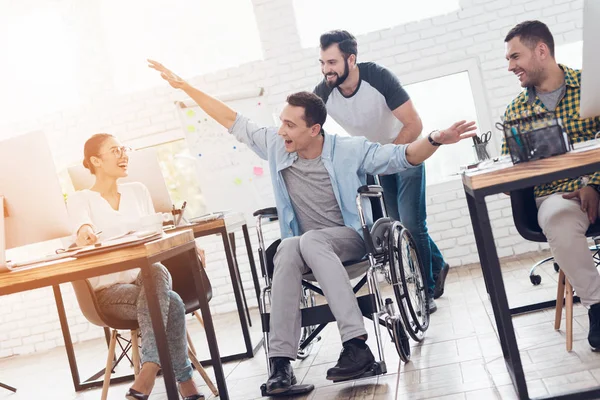 Um colega rola uma pessoa em uma cadeira de rodas ao redor do escritório. Eles se divertem e riem . — Fotografia de Stock