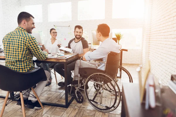 Um homem em uma cadeira de rodas se comunica alegremente com os funcionários do escritório durante uma reunião de negócios . — Fotografia de Stock