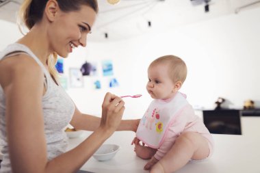 Anne bebek bir kaşık ile beslenir. Çocuk mutfakta masada oturur ve zevkle yiyor.