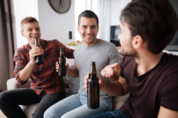 Three men drink beer from dark bottles. They are sitting on the couch and smiling. — Stock Photo, Image