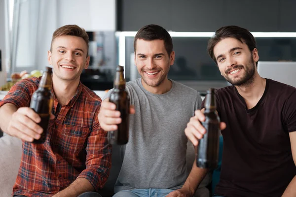 三人从深色瓶子里喝啤酒。他们坐在沙发上, 微笑着. — 图库照片