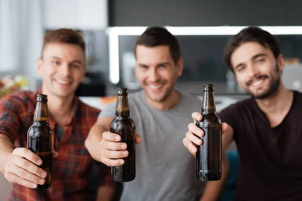 Three men drink beer from dark bottles. They are sitting on the couch and smiling.