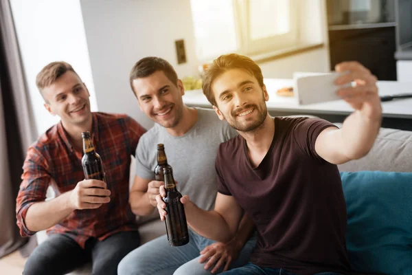 Os homens estão sentados no sofá a beber cerveja. Um homem faz uma selfie com amigos e cerveja . — Fotografia de Stock