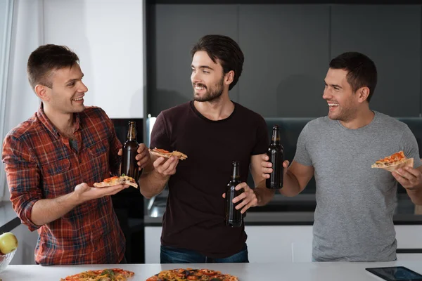 Men drink beer and eat pizza in the kitchen. They talk and have a great time.