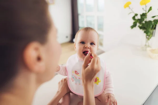 Une femme nourrit un bébé avec une cuillère. Un enfant s'assoit sur une table dans la cuisine et mange avec plaisir . — Photo