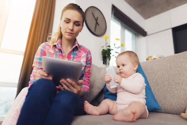 Mama siedzi na kanapie i obok siedzi jej dziecko. Kobieta czyta coś na tablecie. — Zdjęcie stockowe