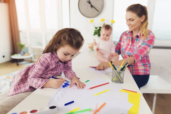 Uma menina desenha na parte superior da cozinha com lápis de cor. A mãe de uma menina com um bebê está sentada lado a lado . — Fotografia de Stock