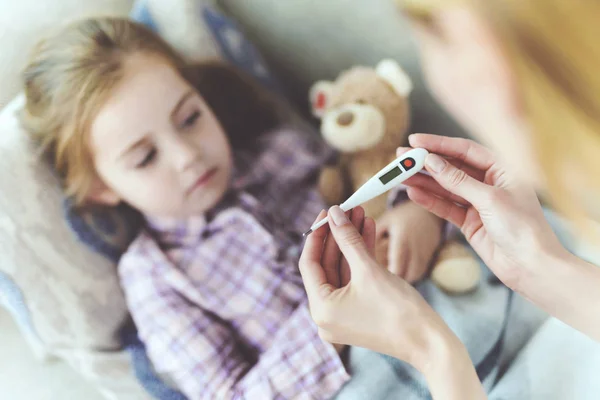 A woman is sitting next to a little girl who is sick. She holds a thermometer, which the girl's temperature measured. — Stock Photo, Image