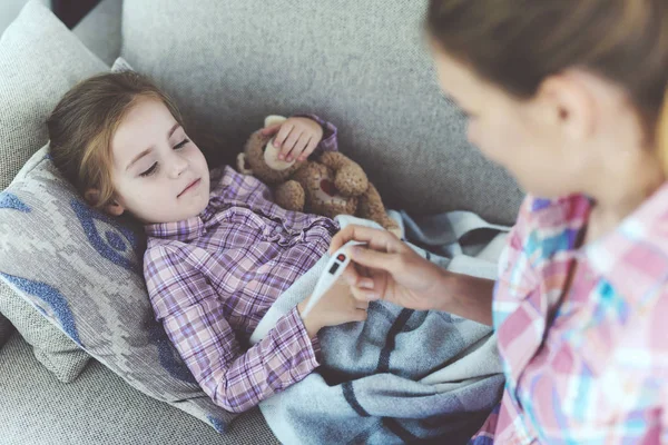 A woman is sitting next to a little girl who is sick. She holds a thermometer, which the girl's temperature measured. — Stock Photo, Image