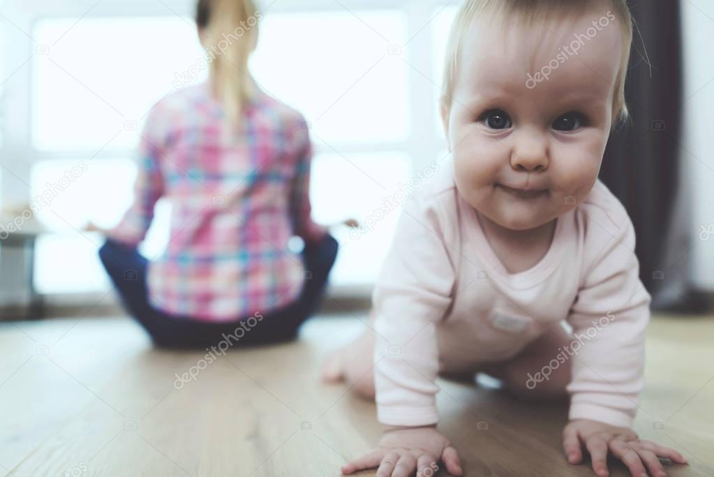 A woman is sitting on the floor and meditating. She does not follow the child who crawls on the floor of the apartment.