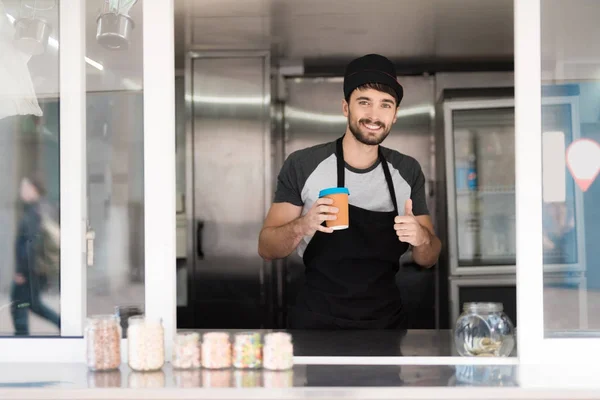 Un hombre en un delantal posando en un restaurante móvil. Él sostiene un vaso de café fragante y muestra un pulgar hacia arriba —  Fotos de Stock