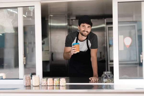 Un hombre en un delantal posando en un restaurante móvil. Él está sosteniendo un vaso de café fragante —  Fotos de Stock