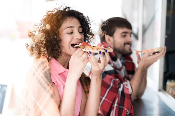 A young couple is standing by a mobile snack bar in the street. They ordered two pieces of tasty pizza and eat it — Stock Photo, Image