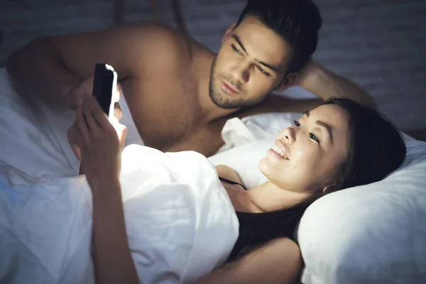 Girl is lying in a dark room in bed with a guy. She looks at the smartphone. Influence of social networks. — Stock Photo, Image