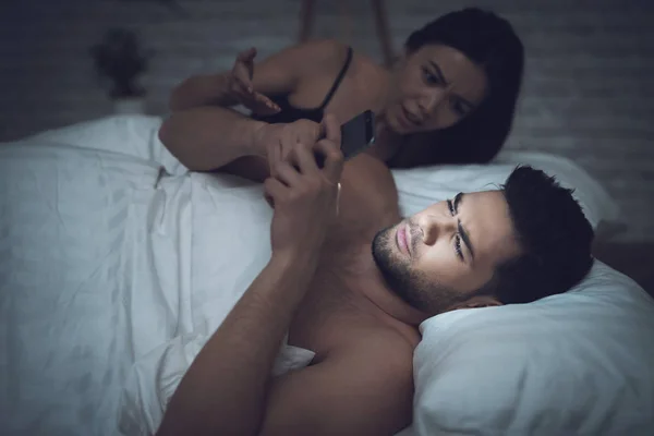 Dziewczyna leży w ciemnym pokoju, w łóżku z mężczyzną. Facet wygląda na smartfon. — Zdjęcie stockowe