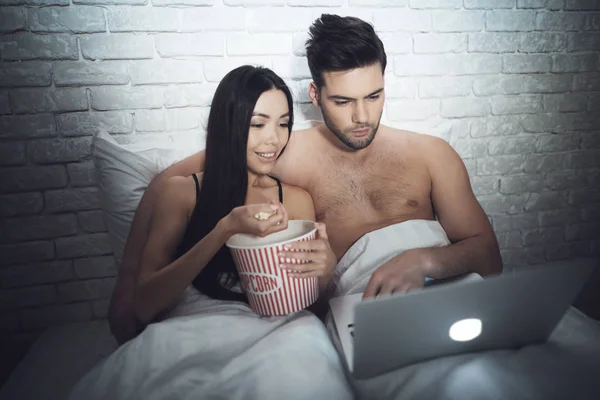 Mädchen liegt in einem dunklen Raum im Bett mit einem Kerl. ein Kerl mit einem Mädchen im Bett schaut auf den Laptop. — Stockfoto