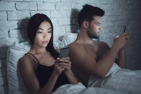 Mädchen mit Kerl liegt in einem dunklen Raum im Bett. Sie schauen auf das Smartphone. Einfluss sozialer Netzwerke. — Stockfoto