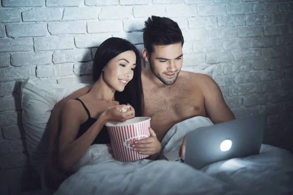 La chica está acostada en un cuarto oscuro en la cama con un chico. Un tipo con una chica en la cama está mirando el portátil . —  Fotos de Stock