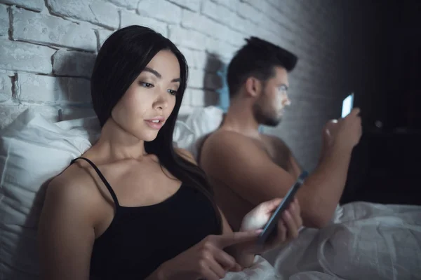 Chica con chico yace en un cuarto oscuro en la cama. Están mirando el teléfono inteligente. Influencia de las redes sociales . —  Fotos de Stock