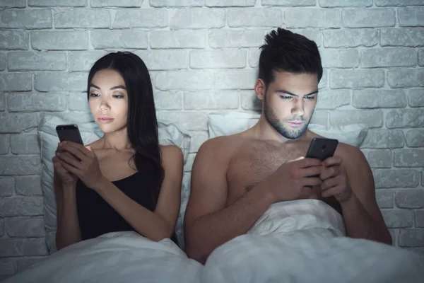 Chica con chico yace en un cuarto oscuro en la cama. Están mirando el teléfono inteligente. Influencia de las redes sociales . —  Fotos de Stock