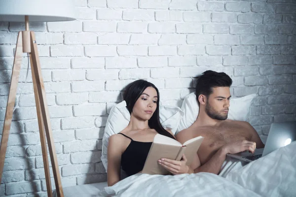 Girl and guy are in bed. A girl reads a book before going to bed. The guy is looking at the laptop.