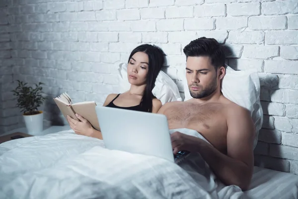 Girl and guy are in bed. A girl reads a book before going to bed. The guy is looking at the laptop.