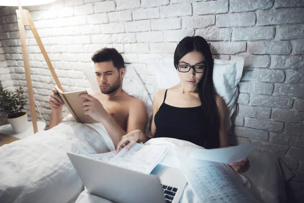 Mädchen und Mann liegen im Bett. liest der Kerl das Buch, bevor er zum Sprung ansetzt. Mädchen arbeitet am Laptop im Bett. — Stockfoto