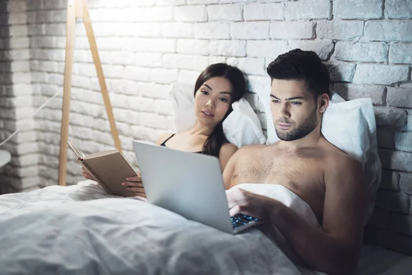 Chica y chico están en la cama. Una chica lee un libro antes de acostarse. El tipo está mirando el portátil. . —  Fotos de Stock