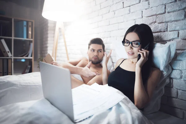 Chica y chico están en la cama. El tipo lee el libro antes de irse a dormir. Chica trabajando en el ordenador portátil en la cama . —  Fotos de Stock