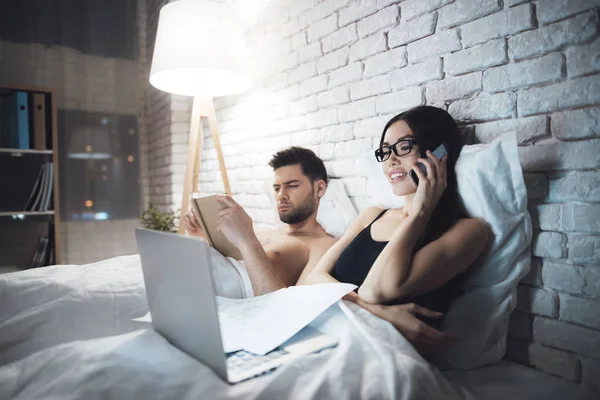 Chica y chico están en la cama. El tipo lee el libro antes de irse a dormir. Chica trabajando en el ordenador portátil en la cama . —  Fotos de Stock