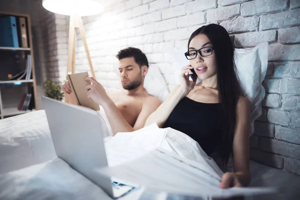 Mädchen und Mann liegen im Bett. liest der Kerl das Buch, bevor er zu Bett geht. Mädchen arbeitet am Laptop im Bett. — Stockfoto