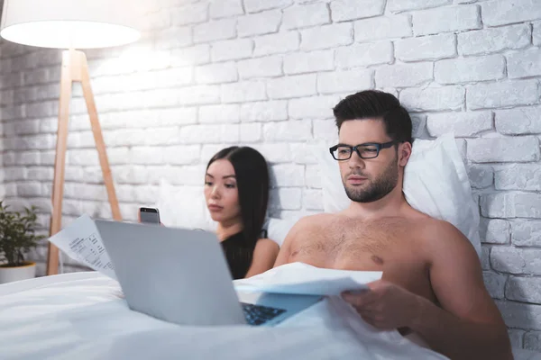 男はベッドに横たわって、ラップトップに取り組んでいます。男は彼のガール フレンドに注意を払っていません。. — ストック写真