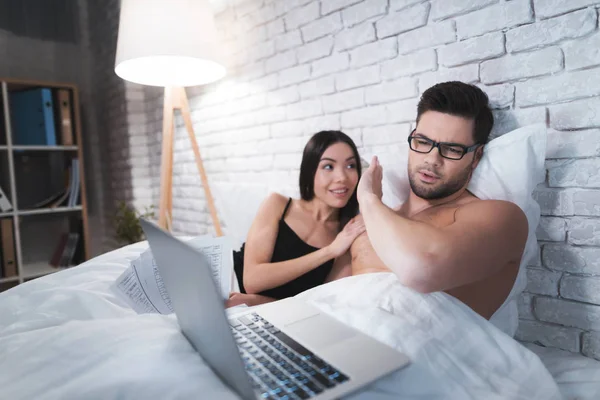 A guy is lying in bed and working on a laptop. The guy does not pay attention to his girlfriend. — Stock Photo, Image