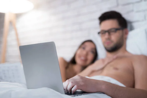 Un ragazzo è sdraiato a letto e lavora su un computer portatile. La ragazza giace sul petto dell'uomo . — Foto Stock