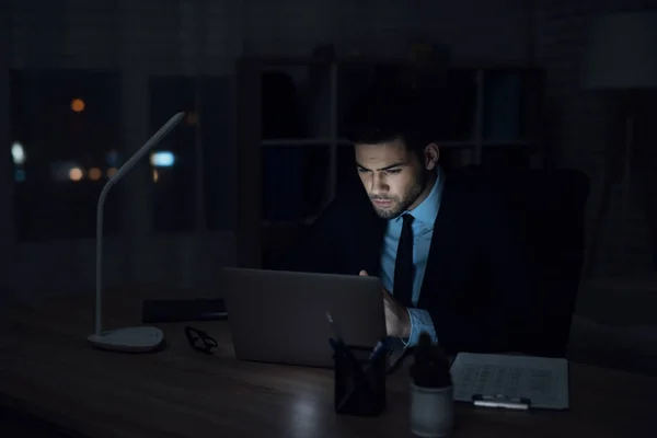 Un uomo e 'seduto davanti a un portatile in un ufficio buio. L'uomo con la tuta lavora . — Foto Stock