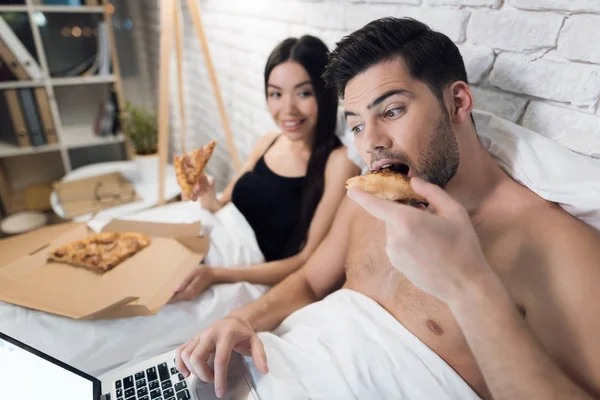 El tipo está acostado en la cama y trabajando en el portátil. Chica está sentada cerca y están comiendo pizza en la cama . —  Fotos de Stock