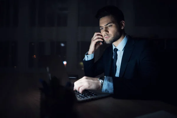 Un uomo e 'seduto davanti a un portatile in un ufficio buio. L'uomo vestito lavora fino a tardi. . — Foto Stock