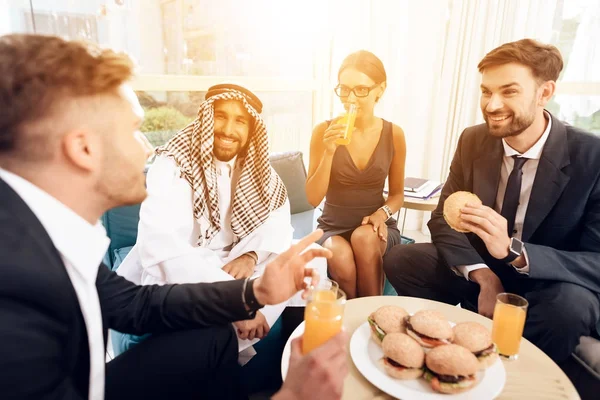 Um homem de roupas árabes e outros homens de negócios estão sentados em uma mesa com hambúrgueres e suco . — Fotografia de Stock