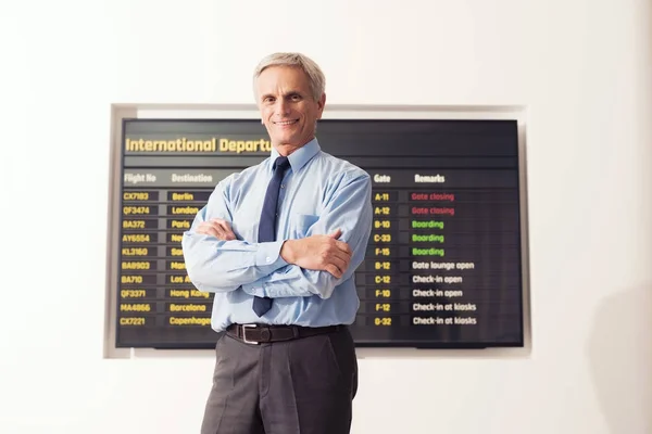 Vanhempi pukumies seisoo lentokentällä. Hänen takanaan on aikataulu. Mies pitelee matkalaukkua. . — kuvapankkivalokuva