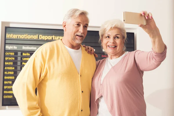 An elderly couple at the airport does selfie. Behind them is the timetable.