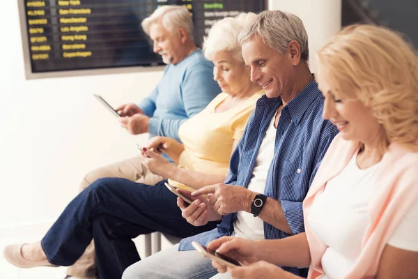 Elderly people are sitting in the waiting room at the airport. They look at their smartphones.