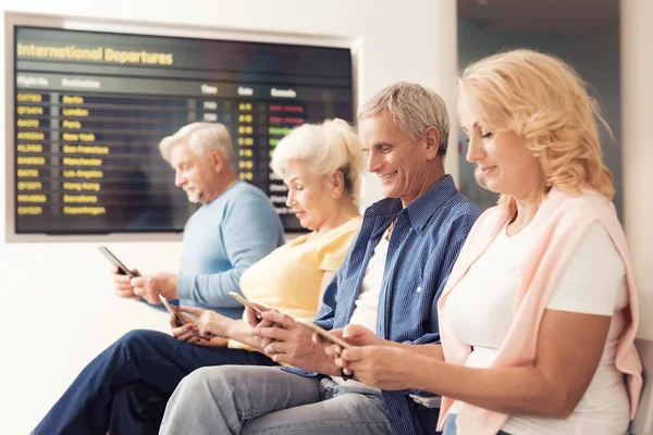 Elderly people are sitting in the waiting room at the airport. They look at their smartphones.