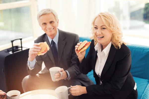 Almuerzo de negocios en la sala del aeropuerto. Un anciano y una anciana en traje comen hamburguesas . —  Fotos de Stock